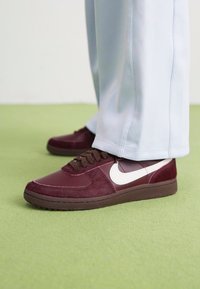 Bordo Nike sportbačiai su tekstūruotais akcentais, balta swoosh ir ruda guma. Pora dėvima su šviesiai mėlynomis kelnėmis ant žalio grindų.