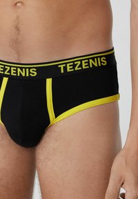 Boxers negros para hombres con acentos amarillos y cinturilla que presenta el nombre de la marca "TEZENIS". La tela parece suave y ajustada.