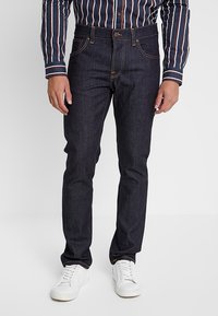 Mörkblå jeans i denim med kontrasterande orange sömmar, raka ben, och knäppning, bärs med vita sneakers och en randig skjorta.