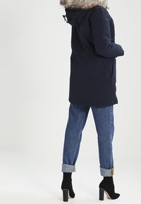 Navy parka med foder av päls, dragkedja och knappstängning, sidofickor och justerbart midjeband. Buren över uppvikta jeans och svarta klackstövlar.