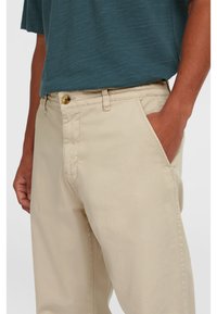 Beige chinos med slim fit, der har en knaplukning, sidelommer og en glat stofstruktur. Båret med en mørkeblå skjorte.