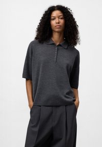 Polo grigio con maniche corte, dotato di colletto e patta con cinque bottoni. Indossato con pantaloni grigi scuri a pieghe. Tessuto liscio.