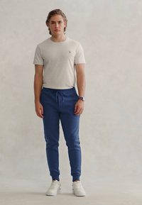 Γκρίζα βαμβακερή μπλούζα συνδυασμένη με μπλε παντελόνια jogger. Τα παντελόνια διαθέτουν κορδόνι με ελαστική μέση και πτυχωμένα πτερύγια. Ένα ζευγάρι λευκά αθλητικά παπούτσια ολοκληρώνει το σύνολο.