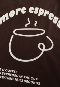 Il tessuto marrone presenta una grafica bianca di una tazza fumante. Il testo sottostante include "18 G Caffè", "Caffè servito in tazza" e dettagli sul tempo di preparazione.