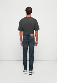 Mann som står med ryggen til, iført en mørkegrå t-skjorte, blå jeans med baketikett og hvite joggesko, mot en enkel hvit bakgrunn.