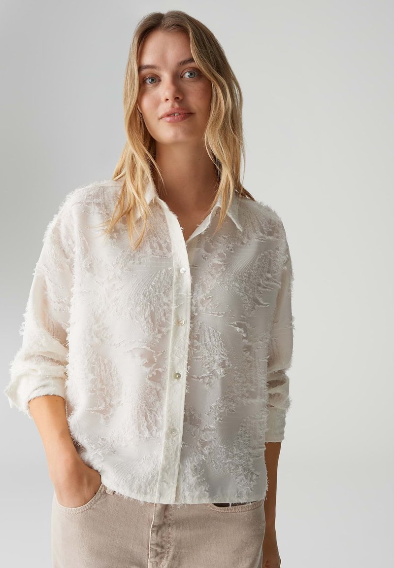 Camicia bianca in tessuto trasparente con motivi strutturati, dotata di chiusura con bottoni sul davanti, maniche lunghe e vestibilità morbida. Tessuto con una leggera lucentezza.