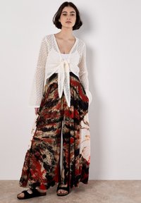 Cardigan blanc en dentelle avec un nœud à l'avant, associé à un pantalon large à motifs en rouge, noir et beige, en tissu léger et fluide.