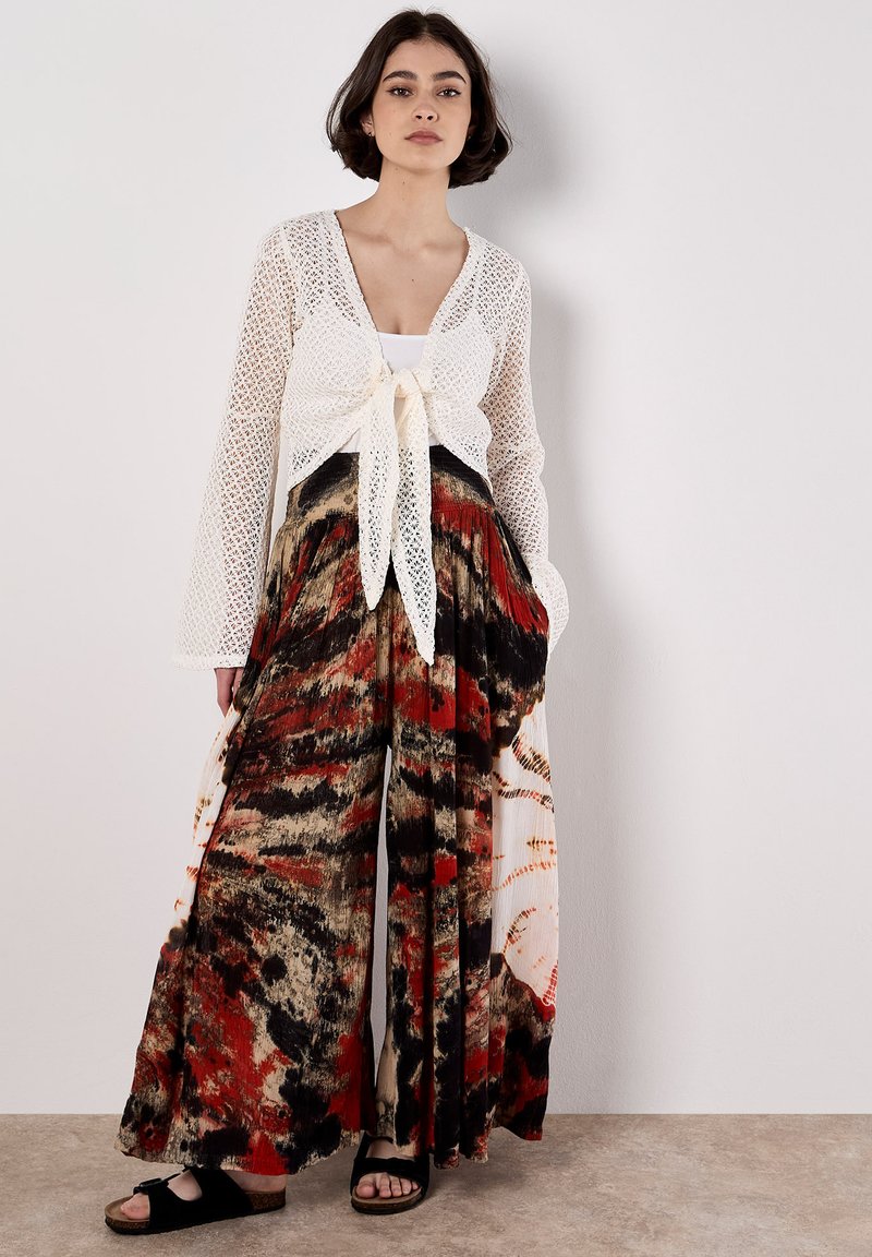 Cardigan blanc en dentelle avec un nœud à l'avant, associé à un pantalon large à motifs en rouge, noir et beige, en tissu léger et fluide.