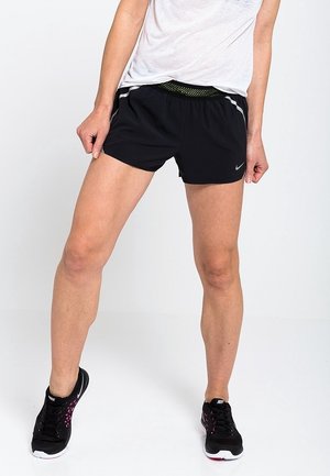 Persoon die zwarte Nike hardloopshorts, een wit los shirt en zwarte Nike sneakers draagt, met gespreide benen staand op een witte achtergrond.