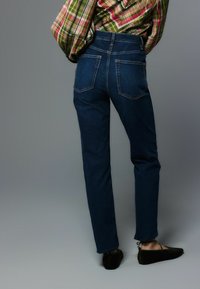 Dunkelblaue Jeans mit figurbetonter Silhouette, dezenten Steppnähten und zwei Gesäßtaschen. Kombiniert mit schwarzen, flachen Schuhen.