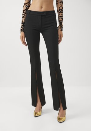 Black tailored pants with long side slits, made of a smooth fabric. Features a classic waist and slim fit, paired with gold high heels.