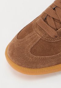 Brązowy zamszowy sneaker z fakturowaną cholewką, zaokrąglonym noskiem i brązowymi sznurówkami, podkreślony podeszwą z gumy.