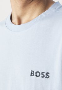 Světle modré bavlněné tričko s žebrovaným kulatým výstřihem. Na přední straně se nachází černé texturované logo "BOSS". Hladká textilie.