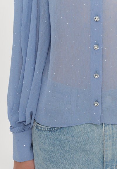 Light blue blouse with long sleeves, textured fabric, sparkling accents, and button closure. Styled with soft cuffs and a relaxed fit.