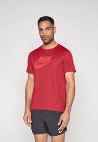 Κόκκινο αθλητικό μπλουζάκι από ελαφρύ ύφασμα, με μεγάλο κόκκινο λογότυπο "RUN" και κλασικό σχέδιο με στρογγυλή λαιμόκοψη. Φορώντας με μαύρο σορτς.