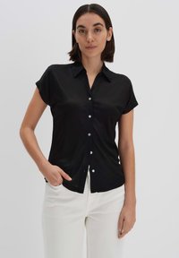 Camicia nera con colletto, maniche corte e una texture liscia. Presenta bottoni di colore chiaro, indossata con pantaloni bianchi.