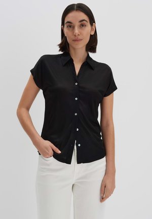 Chemise noire à boutons avec col, à manches courtes, et texture lisse. Présente des boutons de couleur claire, portée avec un pantalon blanc.