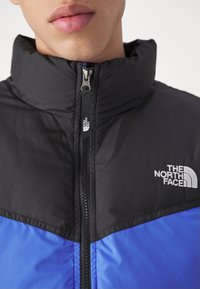 Veste isolante avec une partie supérieure noire et une partie inférieure bleue, col haut, fermeture éclair avant et logo "The North Face" sur la poitrine.