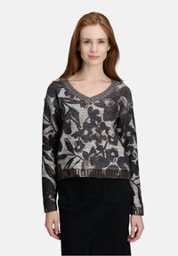 Vrouw met lang rood haar, gekleed in een zwart-wit bloempatronen V-hals trui en een zwarte rok, staand voor een effen achtergrond.