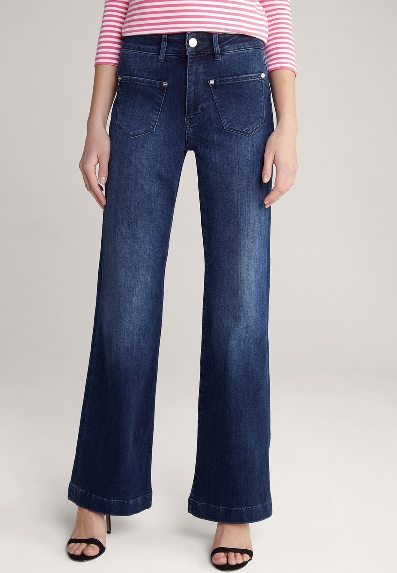 Femme portant un jean bleu foncé à jambes larges, un haut à manches longues rayé rose et blanc, et des talons ouverts noirs, debout devant un fond uni.