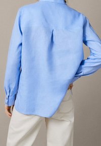 Chemise bleu clair à boutons en tissu léger, avec un pli au dos, des manches longues et des poignets boutonnés, conçue avec un ourlet légèrement incurvé.