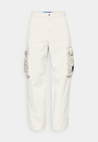 Nákladní kalhoty z lehké béžové bavlny, s dvěma bočními kapsami s klopami, volným střihem a viditelným prošíváním podél švů.