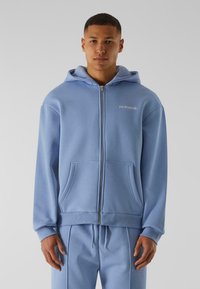 Ljusblå zip-up hoodie i mjukt tyg, med kängurufickor, ribbade muddärmar och huva med dragsko. Diskret logotyp på bröstet.
