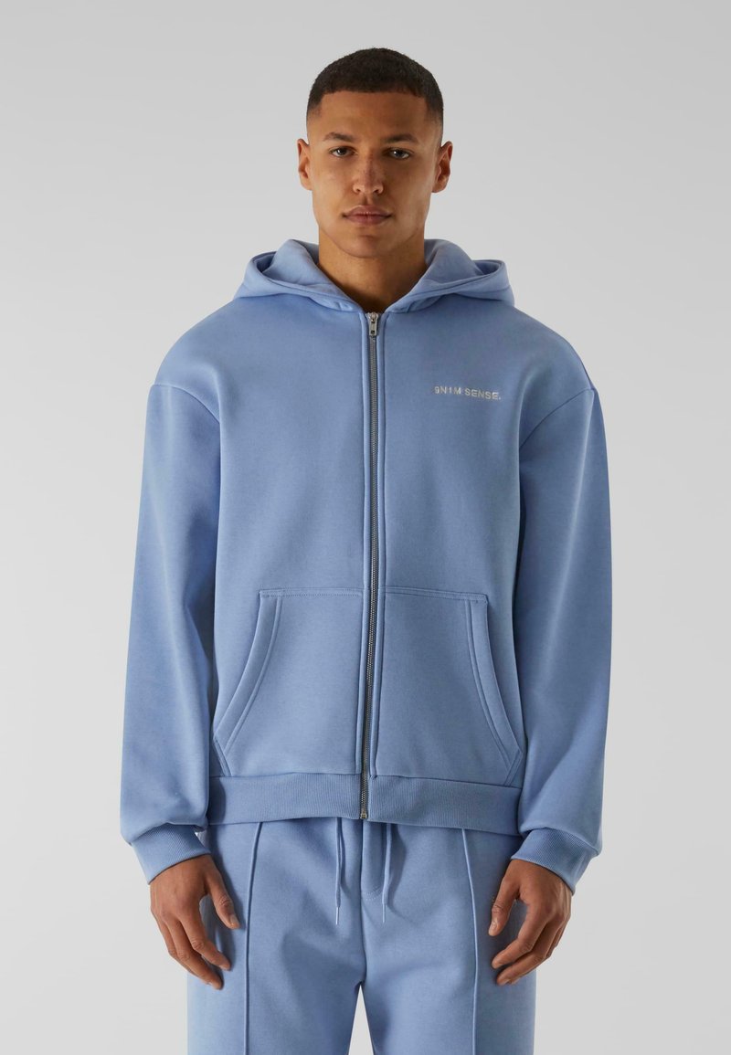Ljusblå zip-up hoodie i mjukt tyg, med kängurufickor, ribbade muddärmar och huva med dragsko. Diskret logotyp på bröstet.
