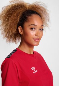 Rød sports t-shirt med v-halsdesign, strukturerede sorte accenter på skuldrene og et hvidt logo på brystet.