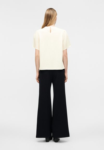 Γυναίκα με μακριά ξανθά μαλλιά φοράει εκρού μπλούζα με κοντά μανίκια και άνοιγμα στην πλάτη, μαζί με μαύρο παντελόνι φαρδιάς πατούσας, στέκεται με την πλάτη στραμμένη.