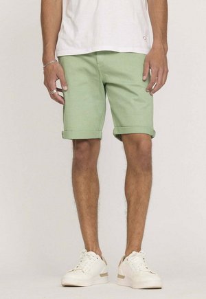 Shorts en coton vert clair avec des revers, associés à des baskets blanches. Les shorts présentent un design simple et une finition texturée propre.
