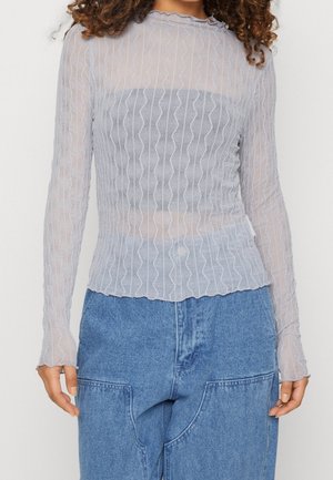 Žena nosící světle šedý průsvitný top s dlouhým rukávem a vlnitými vertikálními proužky přes tmavě šedé tílko, v kombinaci s modrými džínovými kalhotami.