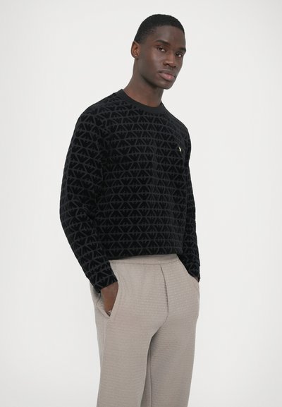 Μαύρο βελούδινο πουλόβερ με υφασμένα γεωμετρικά σχέδια, στρογγυλή λαιμόκοψη και μικρή χρυσή λεπτομέρεια. Συνδυασμένο με ανοιχτό μπεζ παντελόνι με σχέδια.