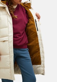 Pufferjack in gebroken wit met een glanzende afwerking, voorzien van een warme bruine voering, ritszakken en een capuchon. Draagt over een bordeauxrode sweatshirt.