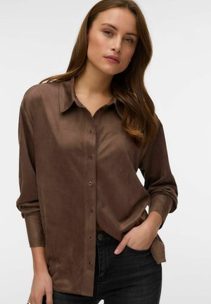 Ung kvinde med langt brunt hår iført en brun skjorte med knapper og sorte jeans, poserer med den ene hånd i lommen mod en ensfarvet baggrund.
