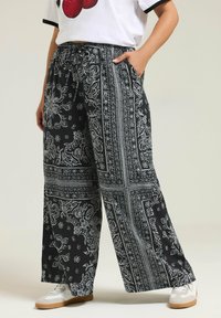 Zwarte wijde broek met een witte intricat bloemen- en paisleydruk, met een elastische tailleband en zijzakken. Vergezeld van witte sneakers.