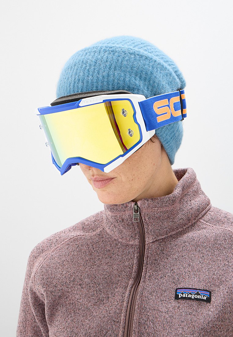 Gafas de esquí con lente reflectante amarilla, marco blanco y correa azul. Modelo que lleva un gorro azul claro y una chaqueta rosa con cremallera.