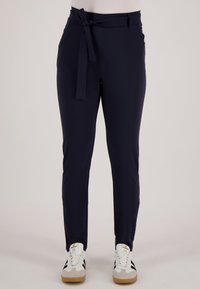 Vrouw draagt marineblauwe taps toelopende broek met riem gestrikt in de taille en witte sneakers met zwarte strepen, staand op een lichte vloer.