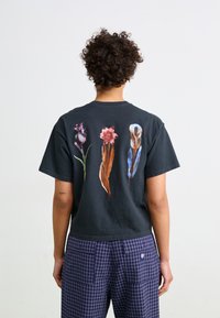 Svart bomull T-skjorte med tre blomsterdesign i nyanser av rosa, grønn og blå trykket på baksiden. Korte ermer, avslappet passform.