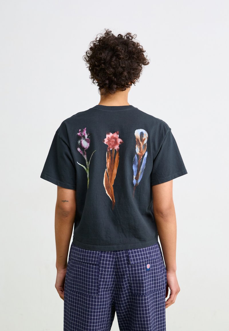 Svart bomull T-skjorte med tre blomsterdesign i nyanser av rosa, grønn og blå trykket på baksiden. Korte ermer, avslappet passform.