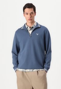 Μπλε πουλόβερ με φερμουάρ, που διαθέτει ριγωτό γιακά. Φορεμένο πάνω από πουκάμισο με καρό σχέδιο, σε συνδυασμό με μπεζ παντελόνι. Λείο υλικό.