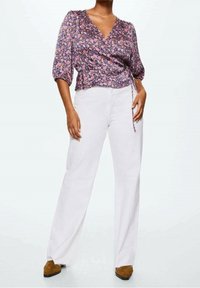 Top floreale avvolgente in tessuto viola, rosa e arancione con maniche a sbuffo; abbinato a pantaloni bianchi a gamba larga e scarpe color cuoio.
