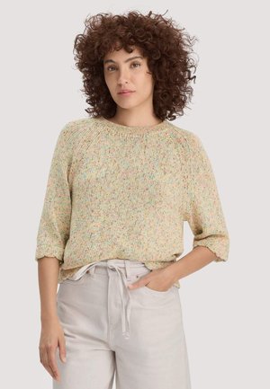 Lockige Frau mit beigem, gesprenkeltem Strickpullover mit hochgekrempelten Ärmeln und hellbeiger, hoch taillierter Hose, die eine Hand in der Tasche hat.