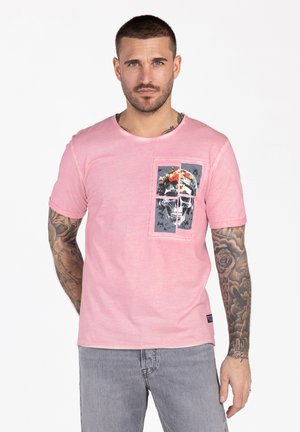 Uomo con capelli corti e tatuaggi sulle maniche, indossa una maglietta rosa con grafica di teschio e fiore nella tasca sul petto, in piedi contro uno sfondo bianco.