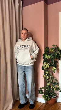 Junge Person in einem hellgrauen Ripcurl-Hoodie und lockeren hellblauen Jeans steht neben einer pinken Wand und einem Vorhang neben einer buntgrünen Topfpflanze.