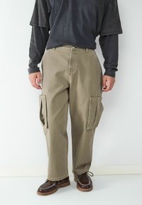 Khaakkleurige katoenen cargobroek met een losse pasvorm, voorzien van zijzakken en zichtbare stiksels. Gecombineerd met een donker langärmige shirt en casual schoenen.