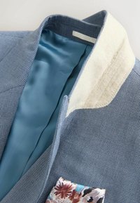 Blazer bleu texturé avec doublure en satin bleu clair, revers de col beige et pochette à motif floral.
