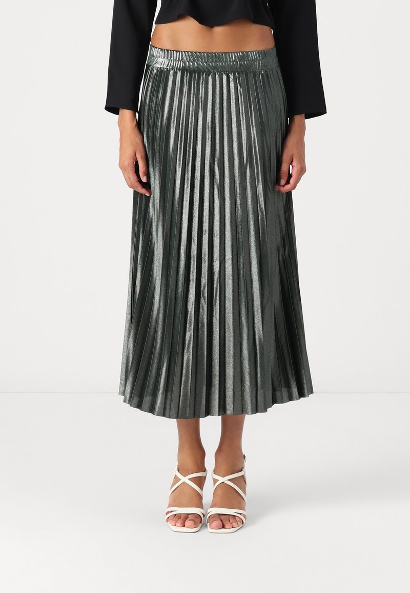 Jupe plissée métallique de couleur vert foncé avec une taille haute. Associée à des talons blancs à lanières. Tissu texturé qui reflète la lumière.
