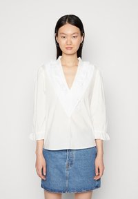 Blusa branca com um colarinho de renda branca com folhos, mangas longas com punhos elásticos e decote em V. Combinada com uma saia de ganga.