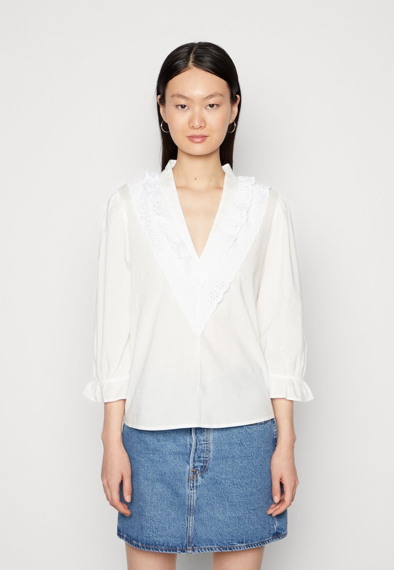 Blusa branca com um colarinho de renda branca com folhos, mangas longas com punhos elásticos e decote em V. Combinada com uma saia de ganga.